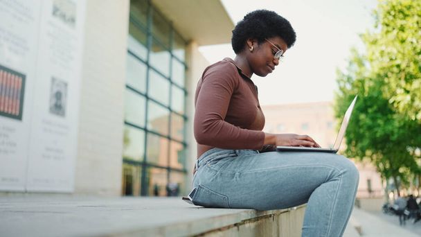 路上でノートパソコンで働く魅力的な女性のフリーランスの側面図。美しいアフロ女性を身に着けているカジュアル勉強屋外 - 写真・画像