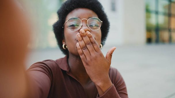 Atraktivní kudrnaté africké ženy posílají vzdušný polibek na kameru, přičemž selfie na ulici, nosí brýle - Fotografie, Obrázek