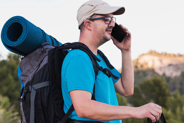 Un excursionista, adulto, varón, caucásico, con una mochila, esterilla, bastones, vestido con una camiseta azul, gorra y pantalones grises con un audífono está en la montaña hablando por su teléfono celular. - Foto, imagen