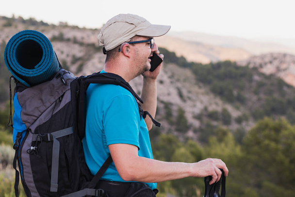 A hiker, adult, male, Caucasian, with a backpack, mat, poles, dressed in a blue T-shirt, cap and gray pants wearing a hearing aid and glasses is on the mountain talking on his cell phone. - Photo, image
