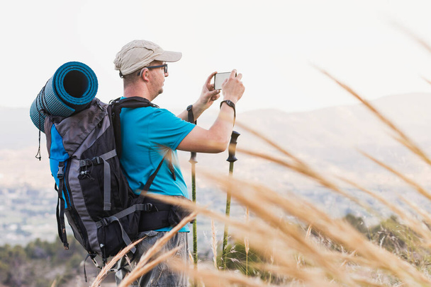 A hiker, adult, male, Caucasian, with a backpack, mat, poles, dressed in a blue T-shirt, cap and gray pants wearing a hearing aid and glasses is on the mountain taking a picture with his cell phone. - Photo, Image