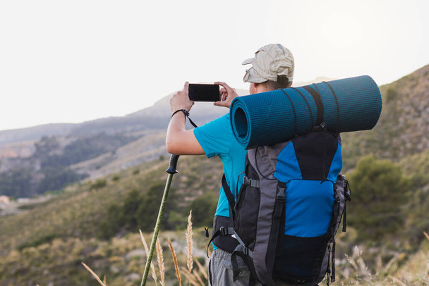 An unrecognizable hiker, adult, male, Caucasian, with a backpack, mat, poles, dressed in a blue T-shirt, cap and gray pants is on the mountain taking a picture of the landscape with his cell phone. - Photo, image