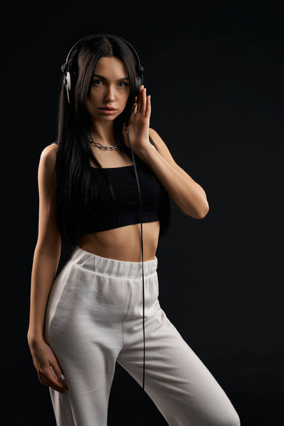 Woman wearing fashion sport clothes listening music with headphones inside. Front view of pretty female model in black top posing with earphones, isolated on black background. Concept of fashion. - 写真・画像