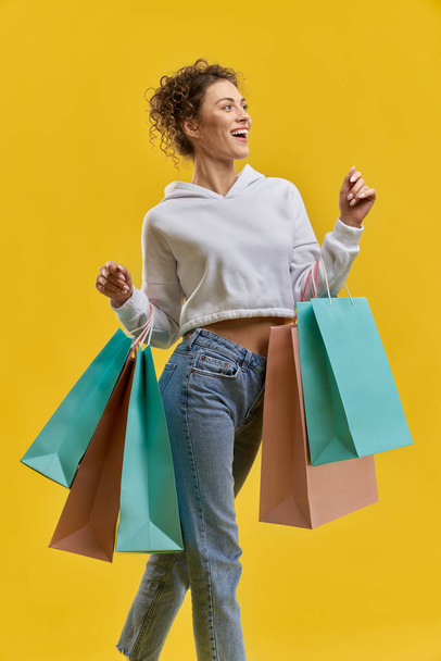 Beautiful girl carrying shopping bags, while looking away in studio. Front view of female shopper with bright bags, walking after shopping, isolated on orange studio background. Concept of shopping.  - Фото, зображення