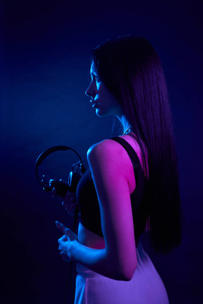 Graceful long haired girl holding headphones, standing straight in dark studio. View from shoulder of brunette female in black top with earphones, highlighted by dark magenta light. Concept of art. - Фото, изображение
