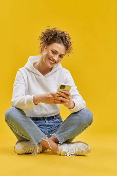 Onnellinen tyttö surffaa internetissä älypuhelimella, kun istuu lattialla. Muotokuva kihara nainen, messaging puhelimessa, istuu ristissä jalat, eristetty oranssi tausta. Sosiaalisen verkon käsite. - Valokuva, kuva