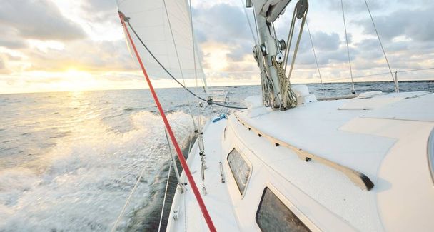 Яхта плаває у відкритому морі на заході сонця. Вид крупним планом на палубу, щогли і вітрила. Чисте небо після дощу, драматичні сяючі хмари, золоте сонячне світло, хвилі та бризки води, циклон. Епічний морський пейзаж
 - Фото, зображення