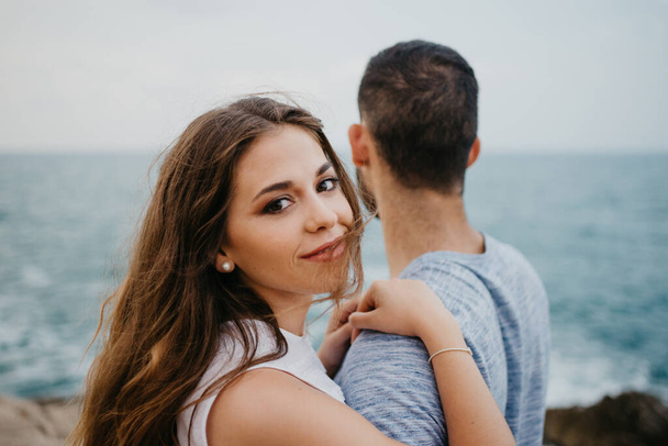 A close portrait of a Latina girl who is keeping her hands on the shoulder of her Hispanic boyfriend on the rocky sea coast in a highland park in Spain. A couple of tourists in the evening in Valencia - Photo, Image