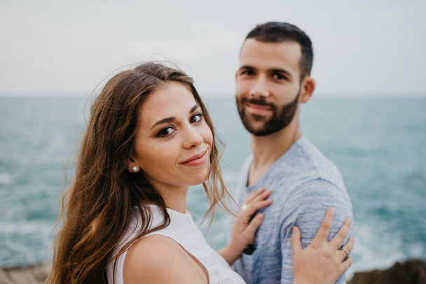A close portrait of a Latina girl who is keeping her hands on the torso of her Hispanic boyfriend on the rocky sea coast in a highland park in Spain. A couple of tourists in the evening in Valencia - Photo, Image