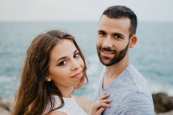 Um retrato próximo de uma menina latina que está mantendo as mãos no tronco de seu namorado sorridente na costa do mar rochoso em um parque de planalto na Espanha. Um par de turistas à noite em Valência - Foto, Imagem