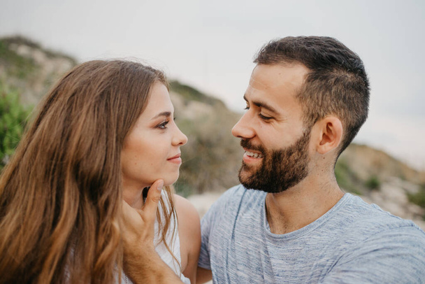 A close photo of a smiling Hispanic man who is stroking with his hands the face of his Latina girlfriend on the rocky sea coast in Spain. A couple of tourists in the evening on the hills in Valencia - Zdjęcie, obraz