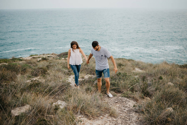 A Hispanic man and his Latina girlfriend are hiking holding hands in a highland park near the sea in Spain. A couple of tourists are enjoying each other on a date at sunset in Valencia. - Photo, image