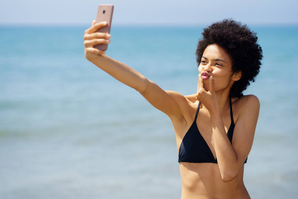 Cheerful African American female traveler in bikini pouting lips and taking self portrait on smartphone near sea in tropical resort - Photo, Image
