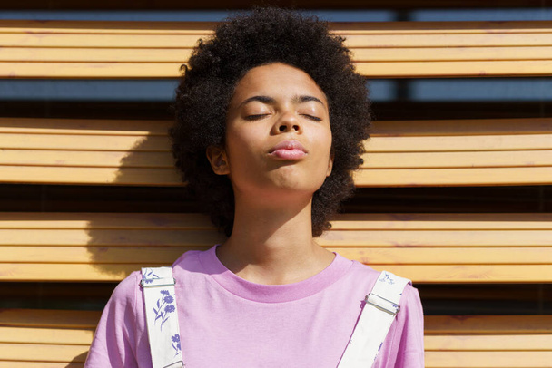 Femme afro-américaine obtenant la lumière du soleil sur son visage avec les yeux fermés, perdu dans la pensée, sur un volet d'un bâtiment moderne. - Photo, image