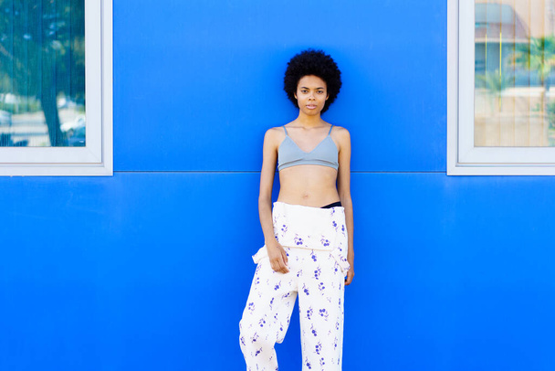 Jovem negra com cabelo afro, modelo de moda, posando contra uma parede azul urbana, vestindo calças e sutiã. - Foto, Imagem