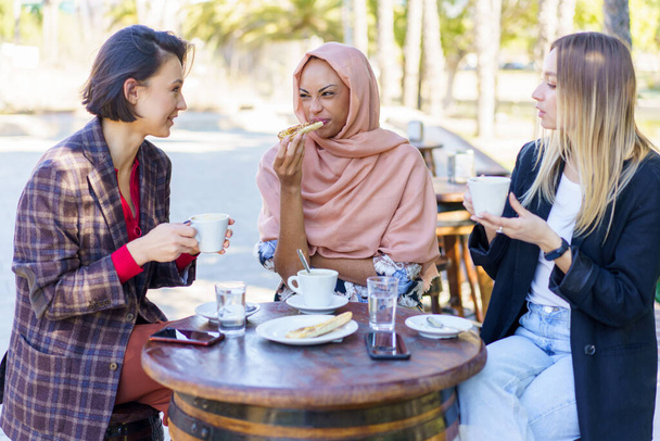 Grupa wielorasowych koleżanek je razem lunch przy stole z filiżankami kawy na tarasie kawiarni na świeżym powietrzu w mieście - Zdjęcie, obraz