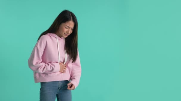 Ziek aziatisch meisje klampen buik terwijl het hebben van stocmach-pijn geïsoleerd op blauwe achtergrond. Ongezonde vrouw die lijdt aan ernstige buikpijn, pijnlijke buikkrampen, indoor studio schot - Video