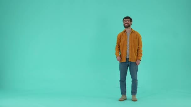 Vrolijke man in bril en casual kleding zwaaien hallo, wijzend vinger naar de camera, tonen uitnodigende gebaar en denkbeeldige verscheidenheid achter hem, terwijl geïsoleerd staan tegen een blauwe studio achtergrond - Video
