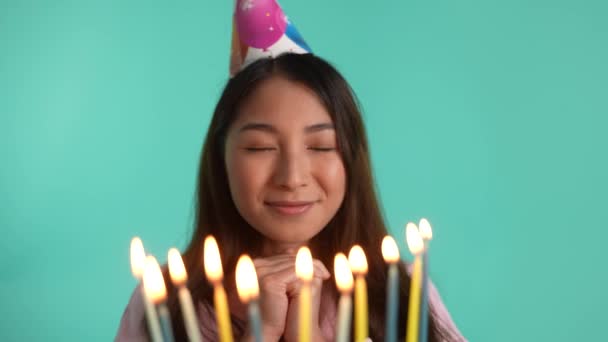 Detailní záběr Narozeninová dívka ve slavnostním kuželovém klobouku přání a sfouknutí svíček izolovaných na modrém pozadí. Veselá asijská fena si užívá narozeninové oslavy, šťastný pocit - Záběry, video