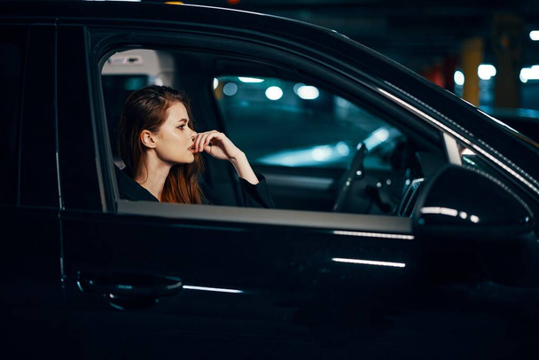 horizontale foto van de zijkant, 's nachts, van een vrouw die in een zwarte auto zit en bedachtzaam vooruit kijkt en haar hand bij haar gezicht houdt. Hoge kwaliteit foto - Foto, afbeelding