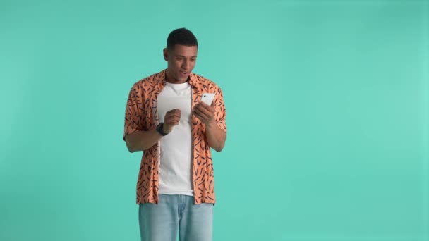 Young dark-skinned man looking at cellphones screen while expecting for result of online betting. Winning man rejoicing, showing yes gesture, expressing success and victory, studio shot - Footage, Video