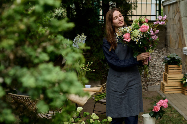portrait of young beautiful florist in apron on terrace making bouquet on green background - Фото, зображення