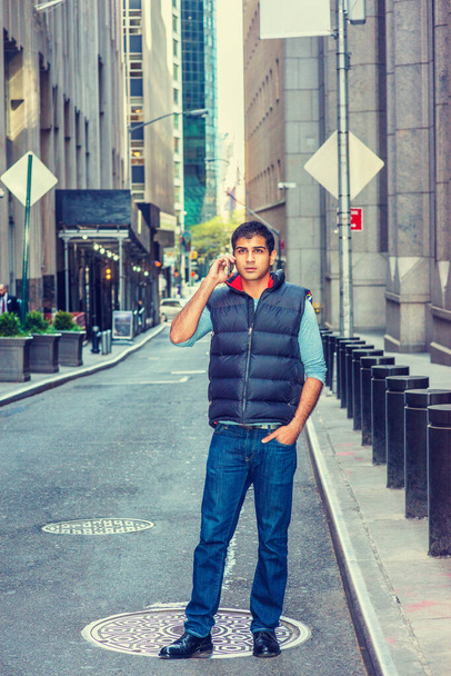 Vida moderna en la ciudad. East Indian American Man con camiseta de manga larga, chaleco azul, jeans, zapatos de cuero, puestos en la estrecha calle vintage de Nueva York, habla por teléfono celular.  - Foto, imagen