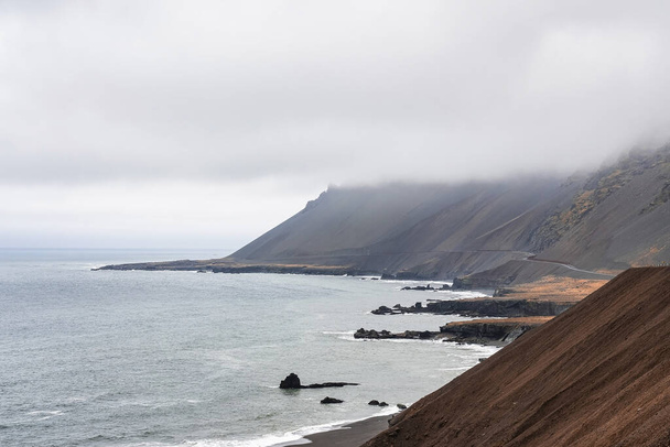 High angle view of steep brown slope. Scenic view of sea waves rushing at black sand beach against cloudy sky. Beautiful scenery of volcanic valley during foggy weather. - Foto, Imagen