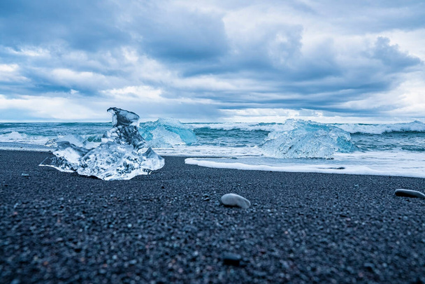 Close-up of beautiful iceberg on black sand at Diamond beach. Chunks of floating ice from Jokulsarlon glacier lagoon against cloudy sky. Scenic view of waves rushing at sea shore. - Foto, Imagem