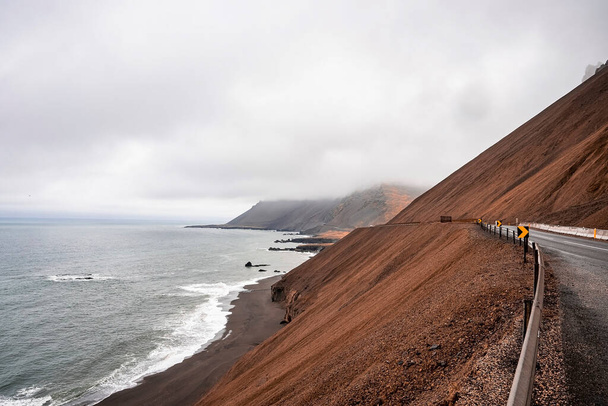 View of steep brown rocky slope beside the ring road. Beautiful sea waves rushing at black sand beach against cloudy sky. Scenic view of volcanic valley during foggy weather. - Fotografie, Obrázek