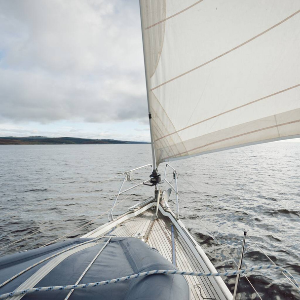 Sloop megbuherálta a modern jachtot fából készült teak fedélzettel, ami a sziklás tó partjainál vitorlázott. Skócia, Egyesült Királyság. Kilátás a fedélzetről az orrra, árbocra, vitorlára. Vándorlás, szabadidős tevékenység, sport, regatta, hajóút - Fotó, kép