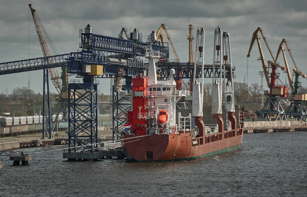 Μεγάλο πλοίο γενικού φορτίου φορτώνει στον τερματικό σταθμό του λιμένα. Ventspils, Λετονία, Βαλτική θάλασσα. Σκοτεινό αστικό τοπίο. Μεταφορές, παγκόσμιες επικοινωνίες, εφοδιαστική, οικονομία, βιομηχανία, περιβάλλον, οικολογικά θέματα - Φωτογραφία, εικόνα