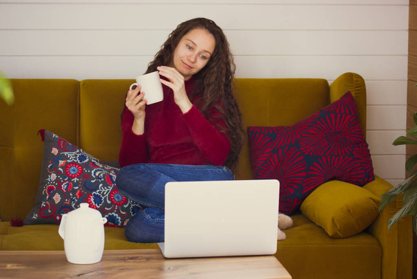Young beautiful Ukrainian woman is sitting on brown couch with laptop on table in cozy home drinking tea from white cup holding in hands watching movie Social media, online communication modern gadget - Φωτογραφία, εικόνα