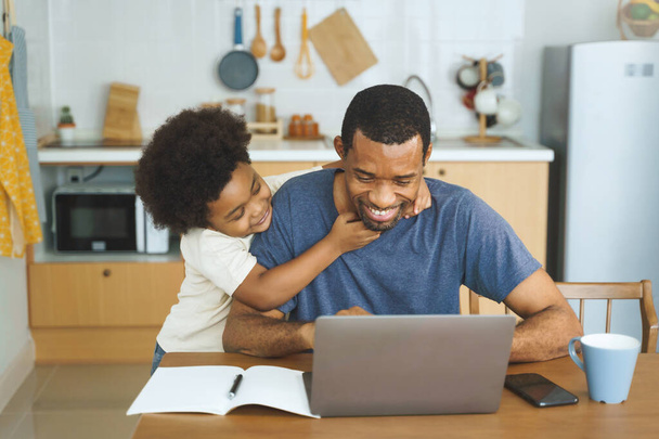 African American father working from home on laptop while his Cheerful son distracts him in kitchen. - Фото, зображення