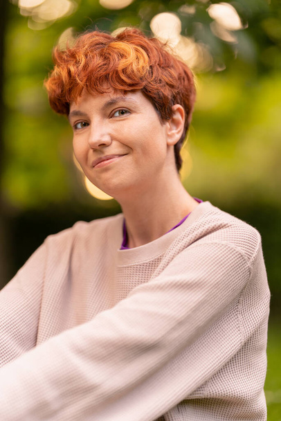 Blij lesbische vrouw in casual sweatshirt met kort roodharig haar glimlachen en kijken naar camera op wazig achtergrond van zomer park - Foto, afbeelding