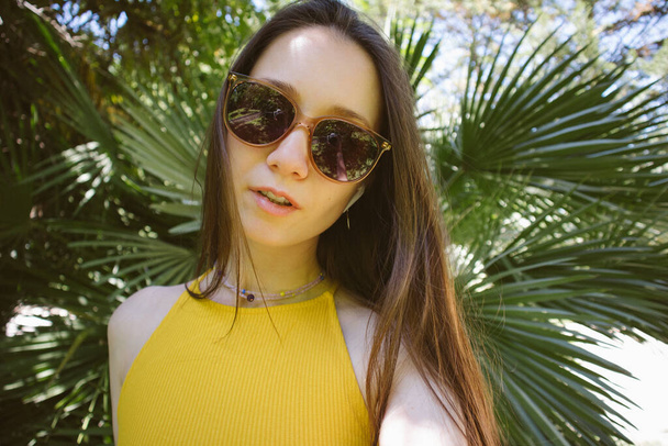 Beautiful young woman, European girl in summer park, garden. Portrait of pretty brunette female solo traveler on green natural background of palm leaves Cute girl in sunglasses yellow dress in tropics - Photo, Image