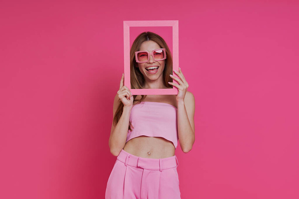 Cheerful young woman looking through a picture frame while standing against pink background - Foto, Imagem