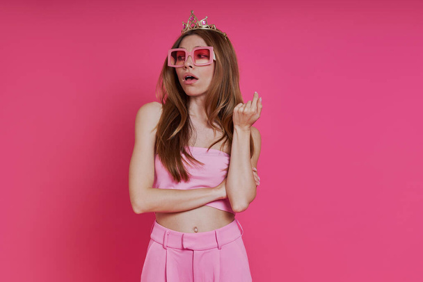 Bossy young woman in funky crown looking displeased while standing against pink background - Fotografie, Obrázek