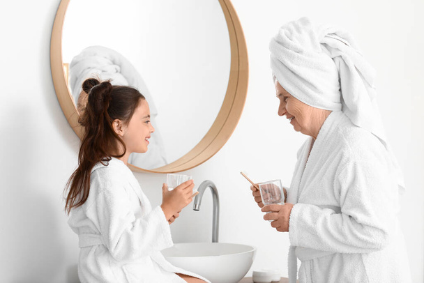 Klein meisje en haar oma met glazen water in de badkamer - Foto, afbeelding