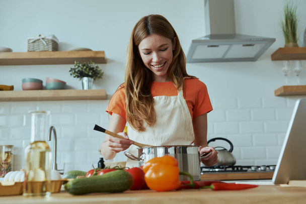 Confident young woman cooking soup while standing at the domestic kitchen - Foto, imagen