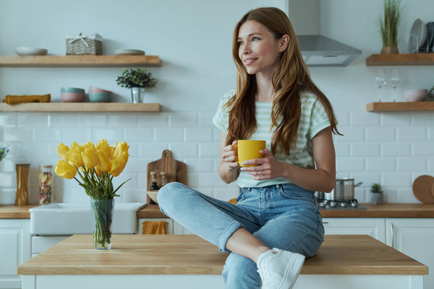 Beautiful young woman holding yellow cup and smiling while sitting on the kitchen counter - Valokuva, kuva