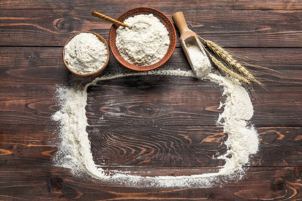 Frame made of scattered flour, bowls, scoop and wheat ears on dark wooden background - Foto, immagini