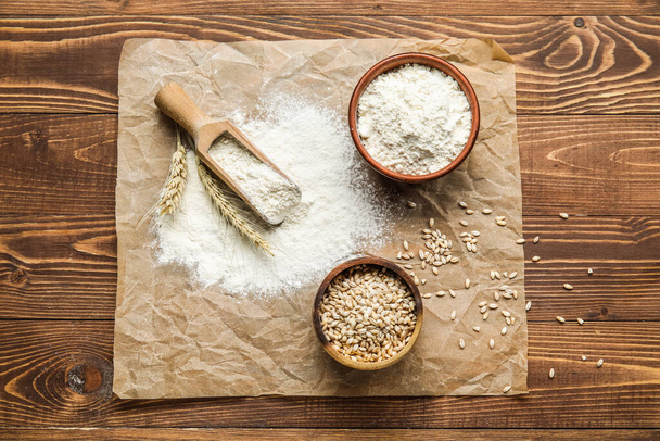 Parchment paper, scoop, bowls with wheat flour and grains on wooden background - Foto, imagen