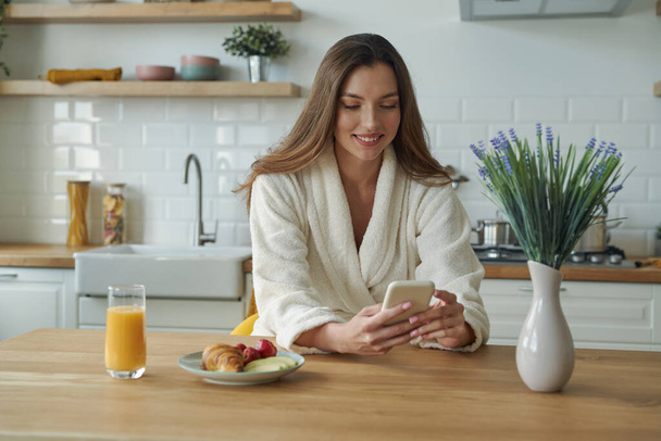 Vonzó fiatal nő fürdőköpenyben okos telefonnal, miközben időt tölt a modern konyhában - Fotó, kép