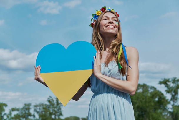 Happy Ukrainian woman in floral crown holding heart shape national flag with sky in the background - 写真・画像