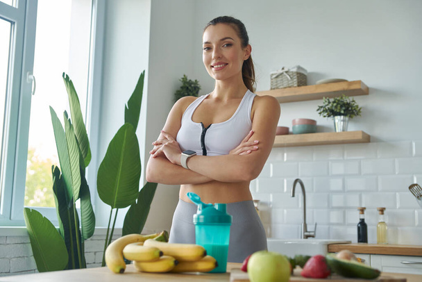 Happy young woman in sports clothing preparing protein cocktail while standing at the kitchen - Foto, Imagem