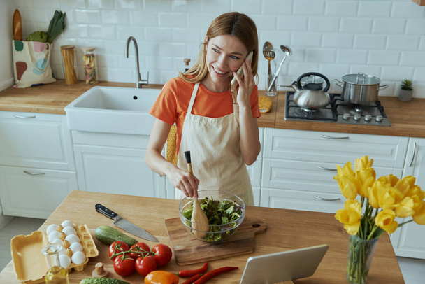 Top view of cheerful young woman talking on mobile phone while cooking at the kitchen - Photo, Image