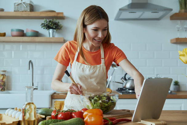 Confident young woman cooking and using digital tablet while standing at the domestic kitchen - Foto, Bild