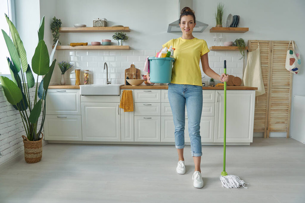 Krásná mladá žena nesoucí mop a čisticí prostředky, zatímco stojí v kuchyni - Fotografie, Obrázek