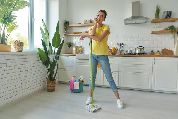 Full length of beautiful young woman carrying mop and smiling while standing at the kitchen - Fotografie, Obrázek